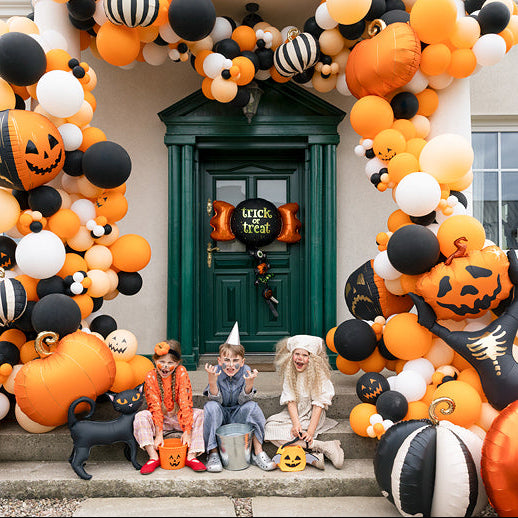 A Halloween decoration setup in front of a house door, featuring a large balloon garland arch in orange, black, and white, pumpkin decorations, a black witch figure, and three children sitting on the doorstep wearing costumes.